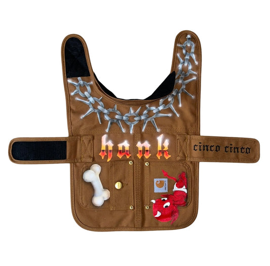 Brown canvas dog vest with hand painted designs on it. A painted barbed wire around the neck, the name hank, the brand cinco cinco, a baby devil and a bone